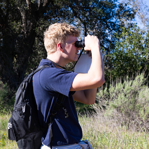 Sideview of a man with blonde hair wearing a navy blue t-shirt and black backpack looks through binoculars while standing outdoors.