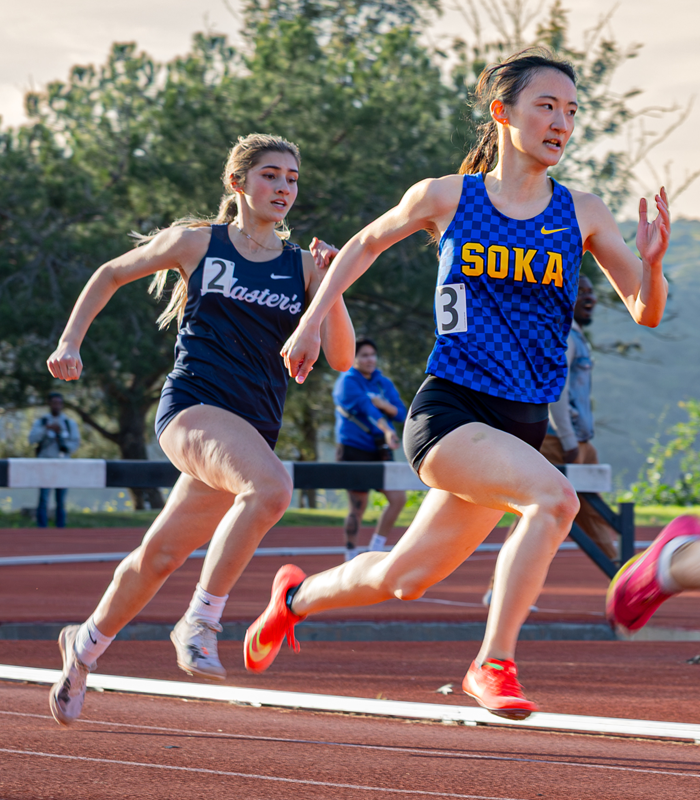 An SUA track athlete wearing a blue tanktop that says SOKA in yellow lettering, runs on a track during a track meet. She is just in front of another runner from a different school.