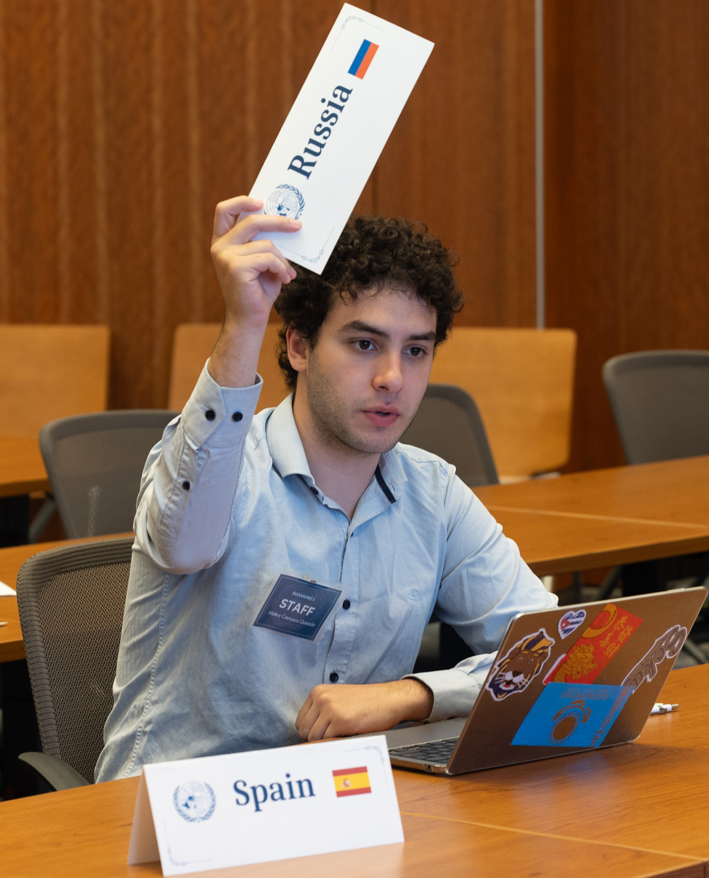 A Soka University Model United Nations team member with curly hair sits at a conference table with a laptop, raising a placard that reads "Russia" while a placard for "Spain" sits on the table in front of him.