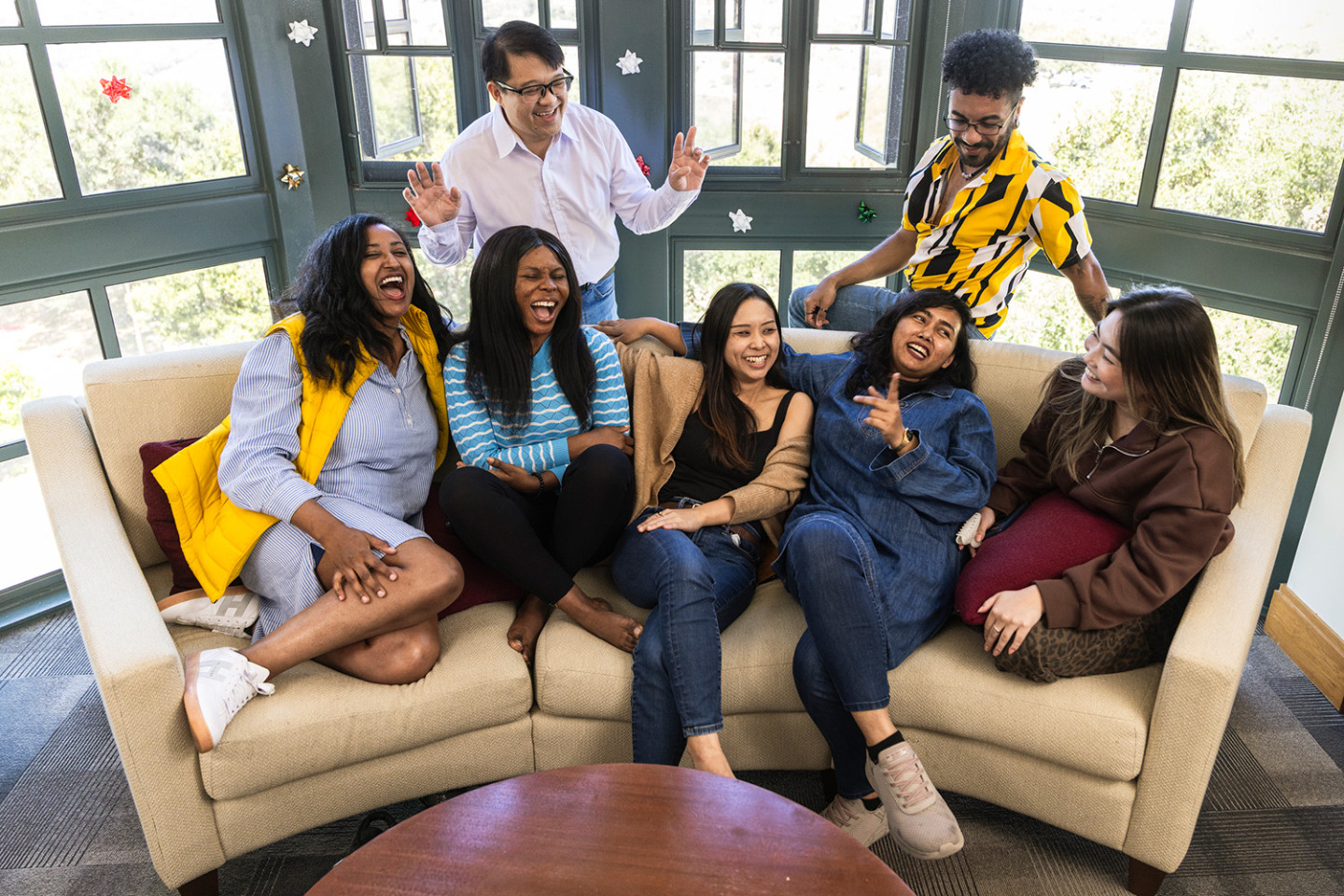 A diverse group of seven friends laughing and hanging out around a curved couch in a sunlit room.