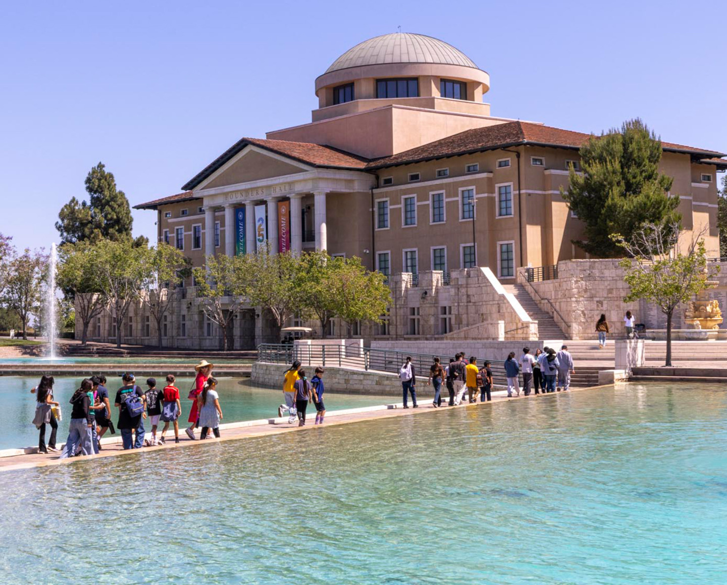 A single-file line of students walking across a low walkway over a large, turquoise reflection pool on a sunny campus, heading toward a large, domed building with a fountain visible in the background.