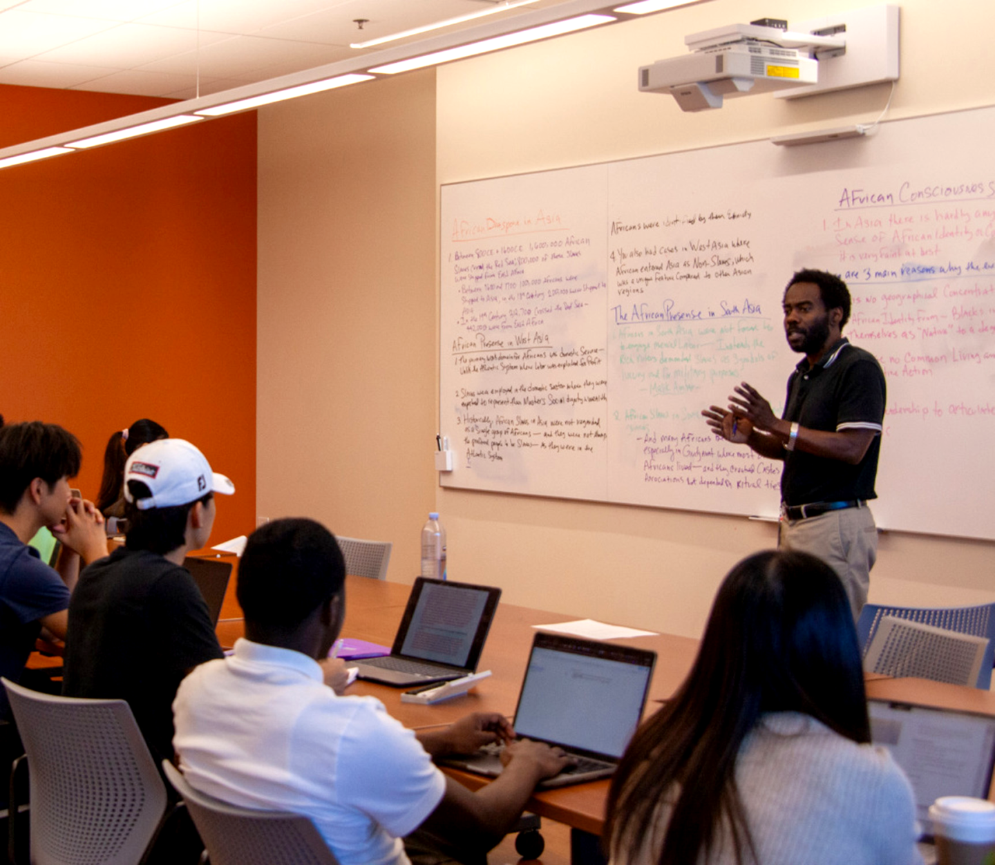 A man stands in front of a white board with a lot of writing on it and gestures with his hands as he speaks to a classroom full of students.