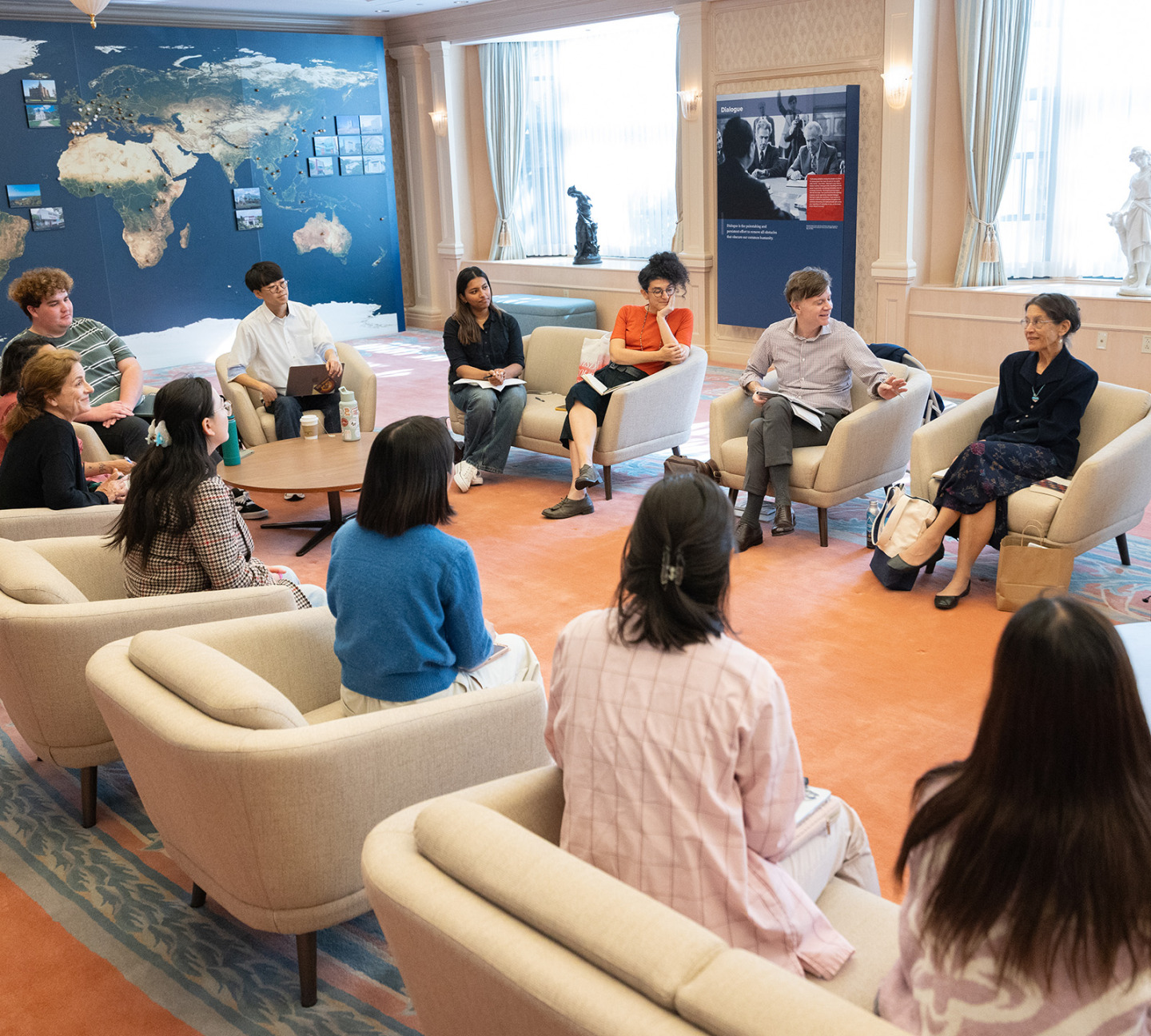 A group of students and professors sit in a large semi-circle of armchairs for a class discussion in a spacious room featuring a large world map mural on the back wall.