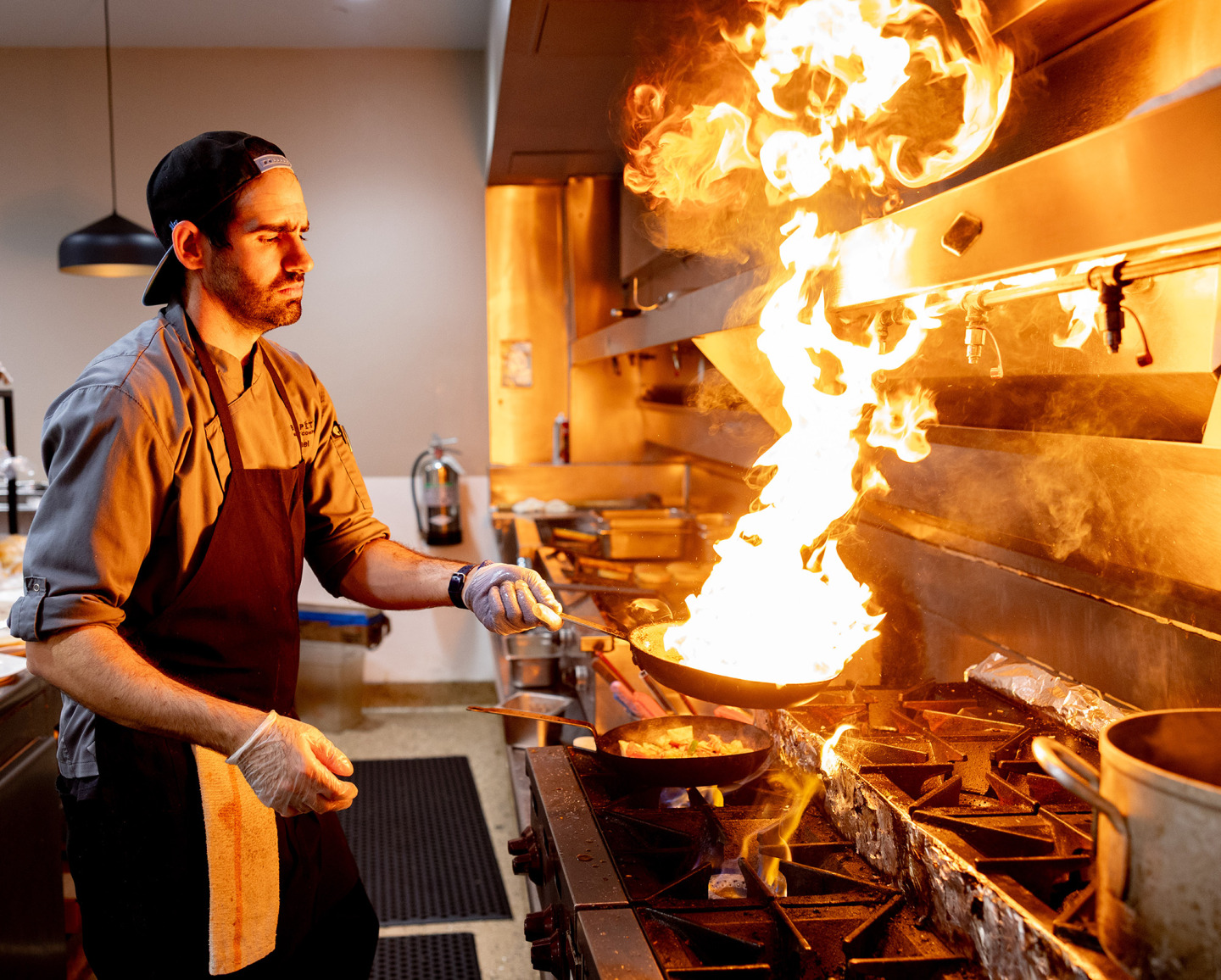 A male chef in a grey uniform and black apron intensely focuses on a large burst of fire erupting from a frying pan on a commercial stove. He wears a backwards baseball cap and gloves while tossing food in a professional kitchen setting.