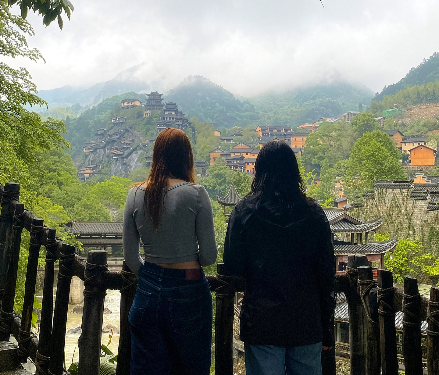  Two women seen from behind look out at a misty mountain village in China featuring traditional wooden stilt houses and structures built into the hillsides.
