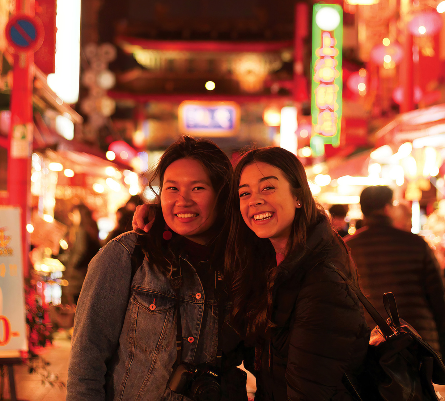 Two young women smiling and posing together at night on a brightly lit street with vibrant red and green neon signs in the background.
