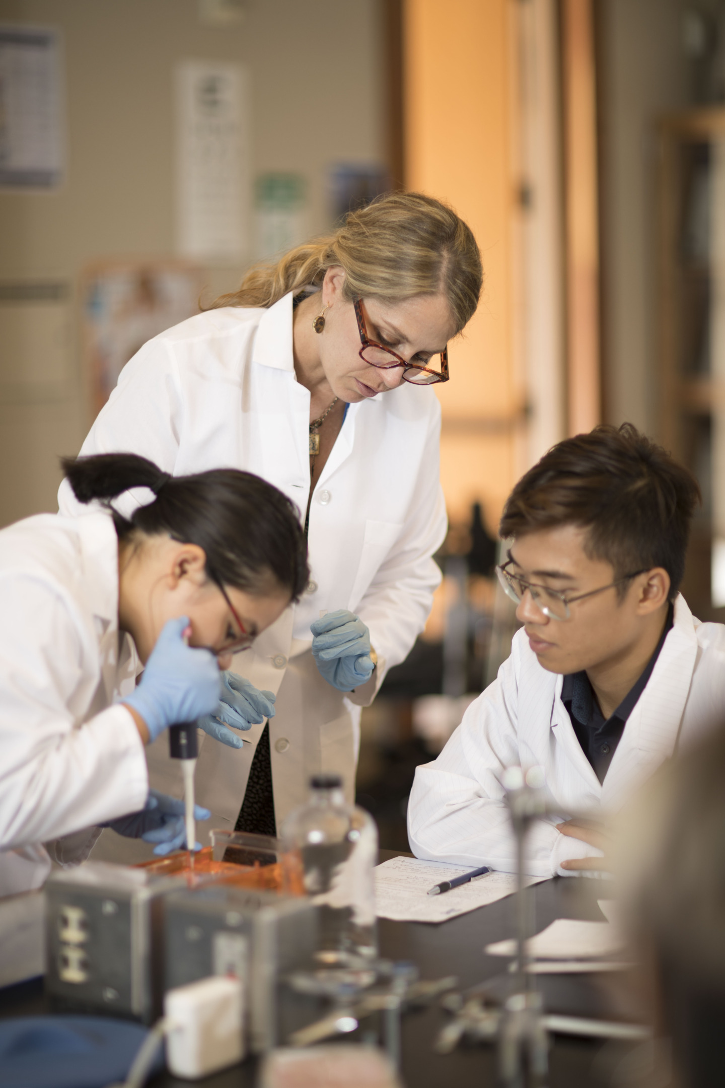 Three people wearing white lab coats and blue gloves perform an experiment in a lab.