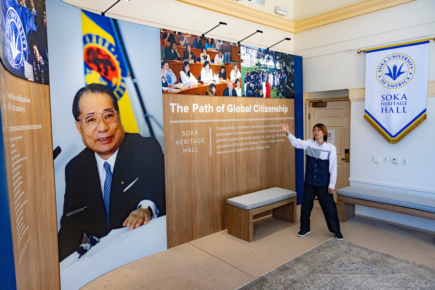 A tour guide points toward an exhibition wall inside the Soka Heritage Hall entry, featuring a large portrait of Daisaku Ikeda and text reading "The Path of Global Citizenship."