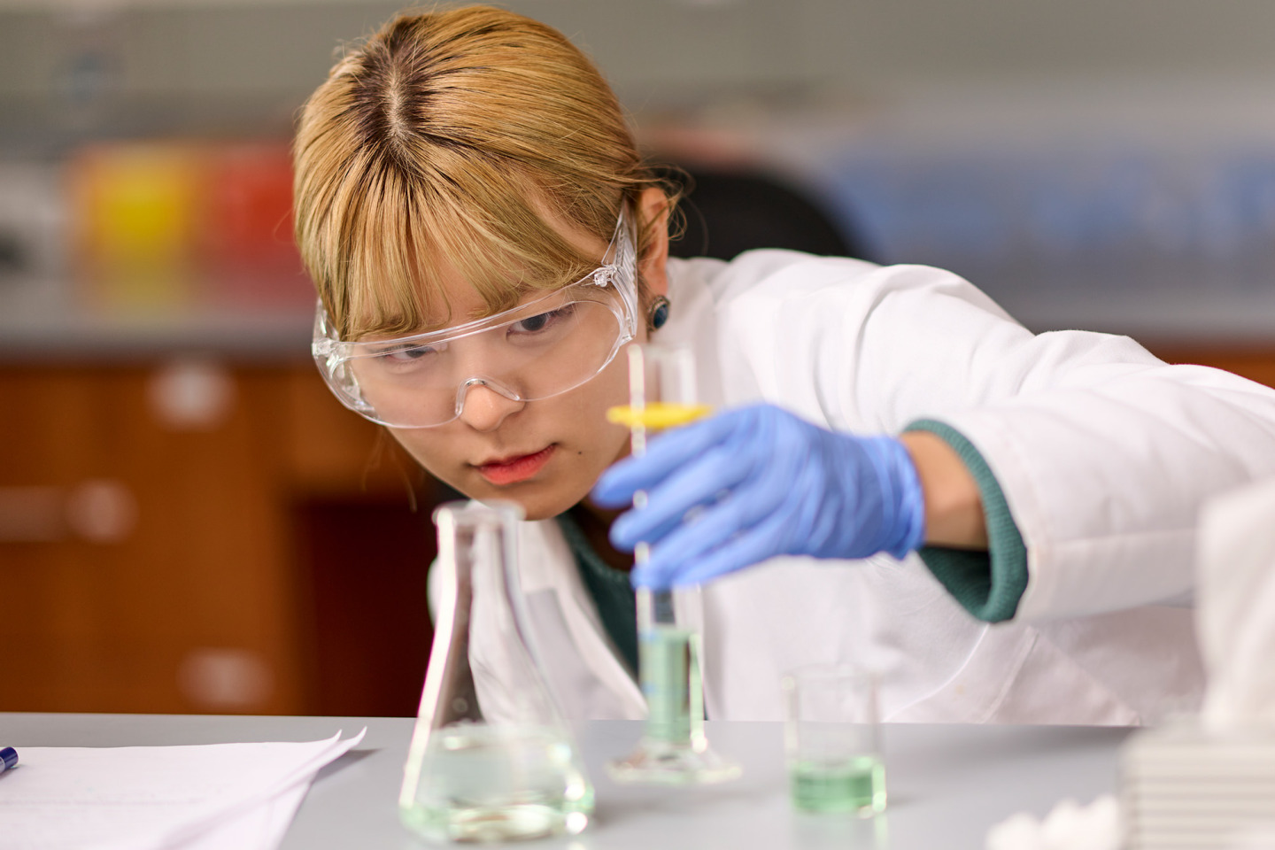 A female student in a white lab coat, safety goggles, and blue gloves leans down to look closely at a graduated cylinder containing a green liquid on a laboratory bench.