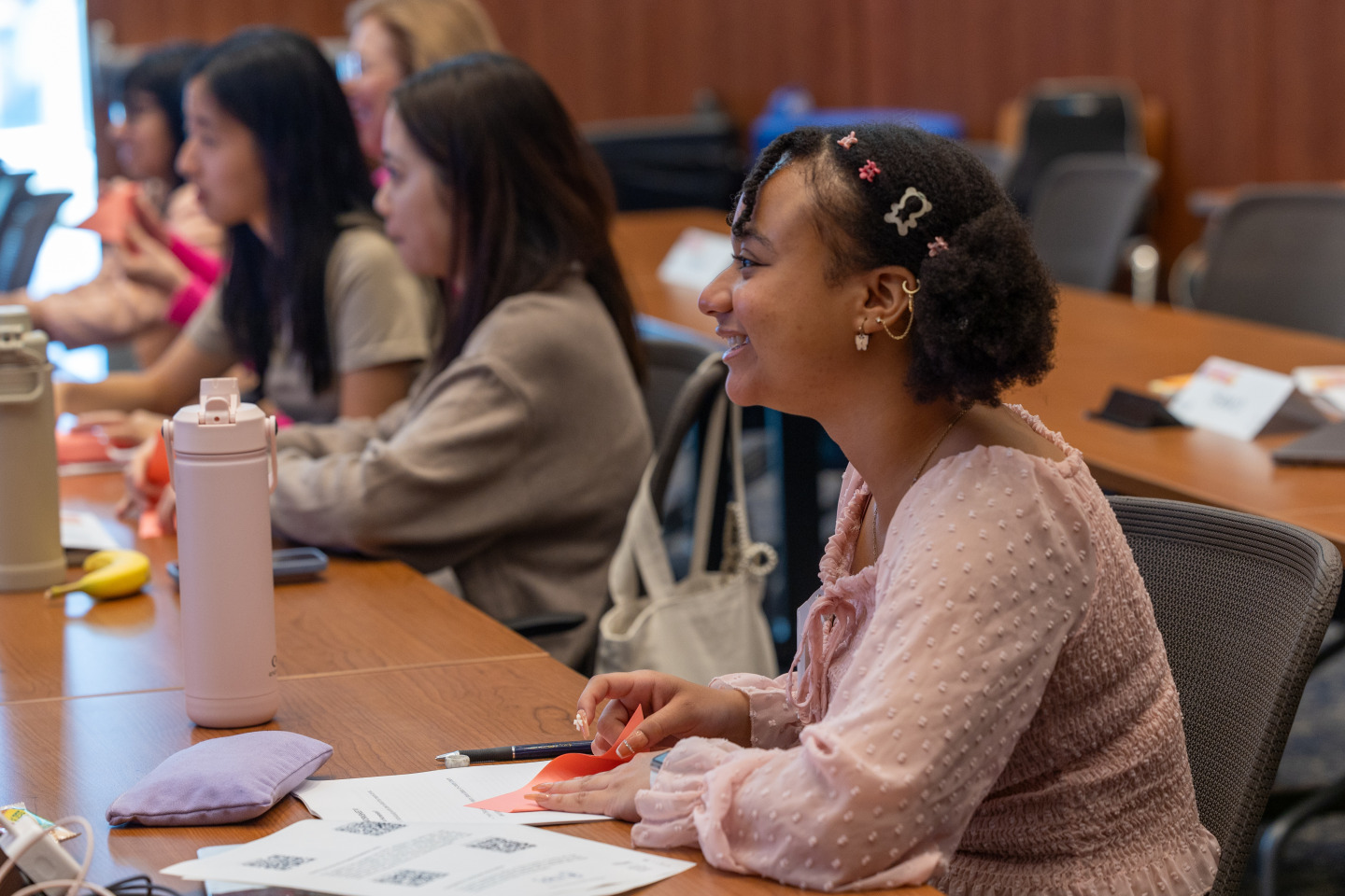 A girl with curly short brown hair wearing a light pink blouse smiles as she listens to a speaker during class.