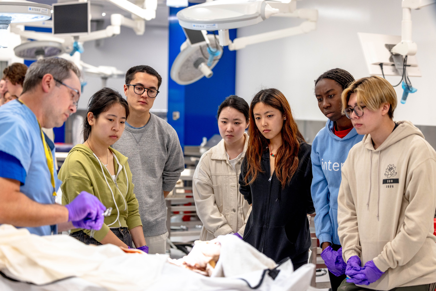 Students look on as a doctor with purple gloves shows them a cadaver during a lab.