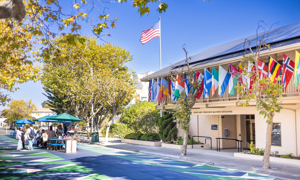 The exterior of a campus building with various country flags hanging over the balcony. A group of people gather on the walkway below.