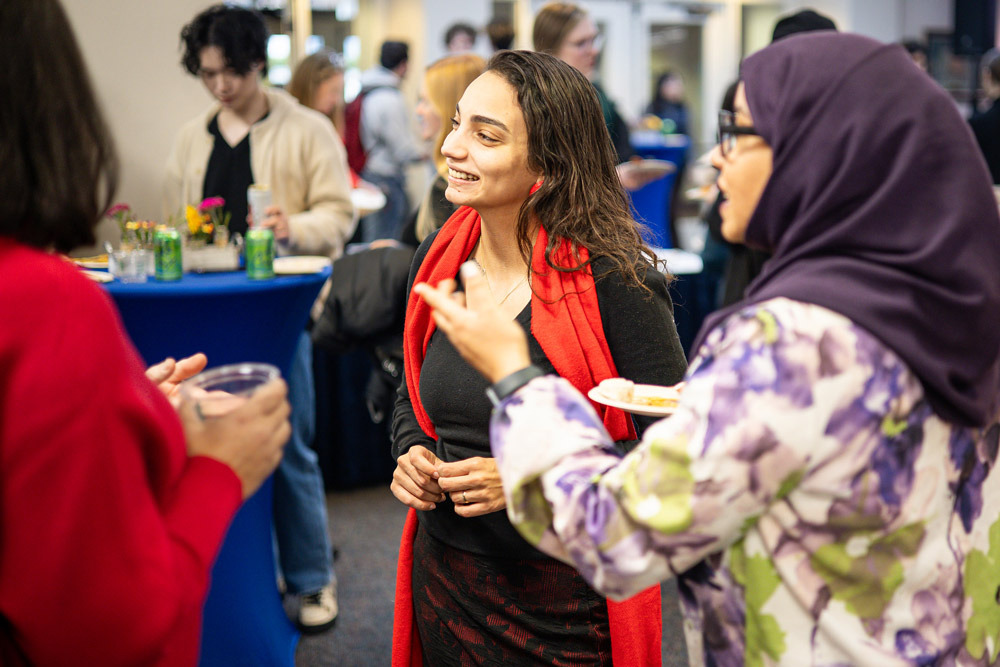 A woman wearing a red scarf over a longsleeve black shirt and red and black pants smiles at someone out of frame. Another person wearing a purple headscarf and green and purple floral shirt points in their direction.