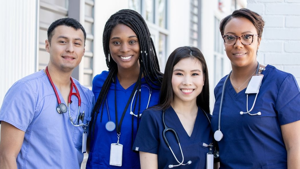 One male nurse stands with 3 female nurses all wearing blue scrubs and stethoscopes around their necks.