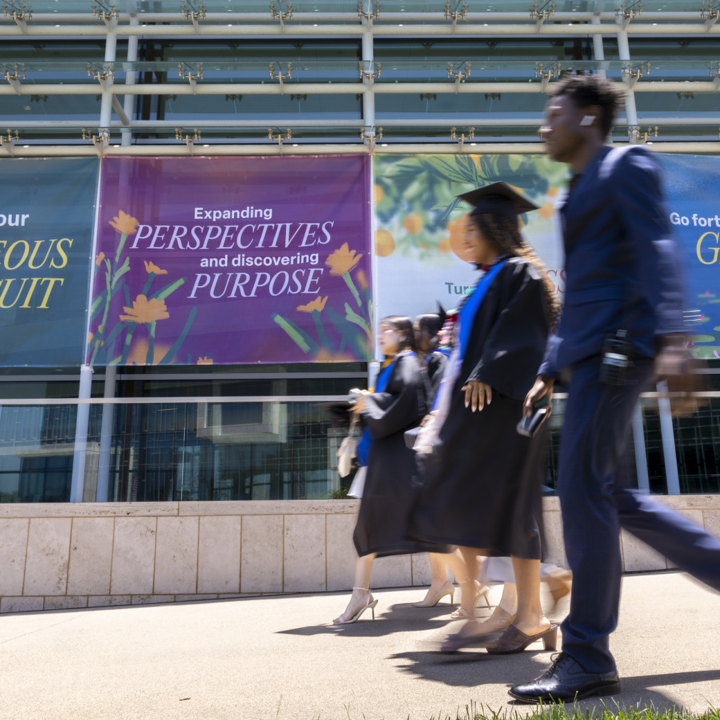 Graduating students walk in a line wearing formal attire and their graduation gowns. They are walking alongside the Performing Arts Center which has branded window clings with mission-oriented language on them.
