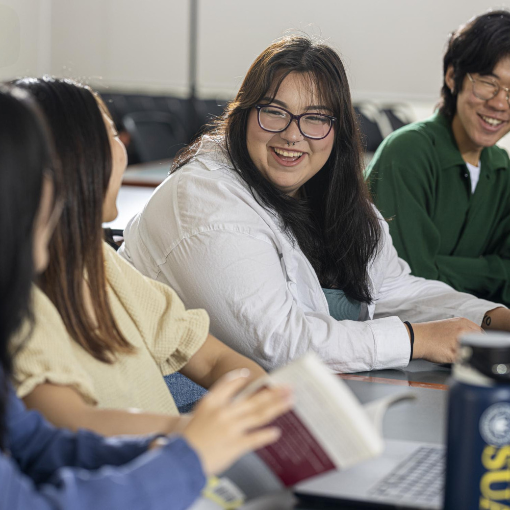 Four students sit in a row facing eachother, smiling and laughing.