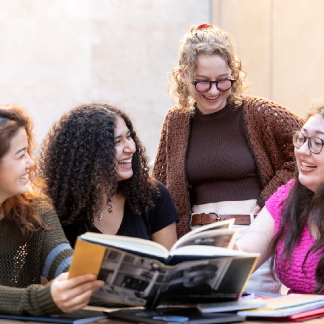 Four young women are gathered around a table outdoors, smiling and laughing together while looking at a large, open book featuring black-and-white photography. One woman stands behind the group, leaning in, while the others are seated.