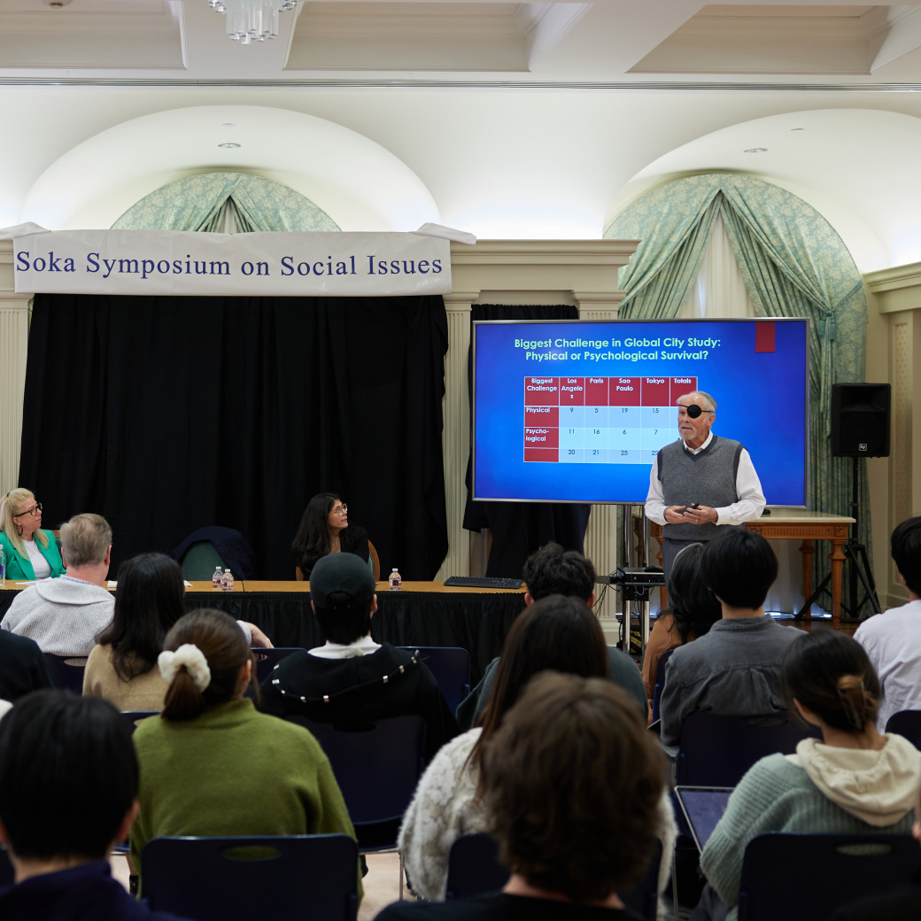 Students sit in rows of chairs and listen to a man with an eyepatch speaking in front of a projector screen. There is a banner overhead that reads, "Soka Symposium on Social Issues." Two women sit in a panel to the side of the man speaking.