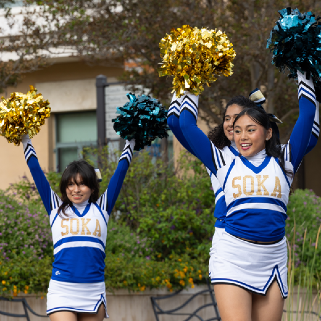Members of Soka's Cheer Student Organization are wearing their cheer uniforms and holding their pompoms above their head during a routine.