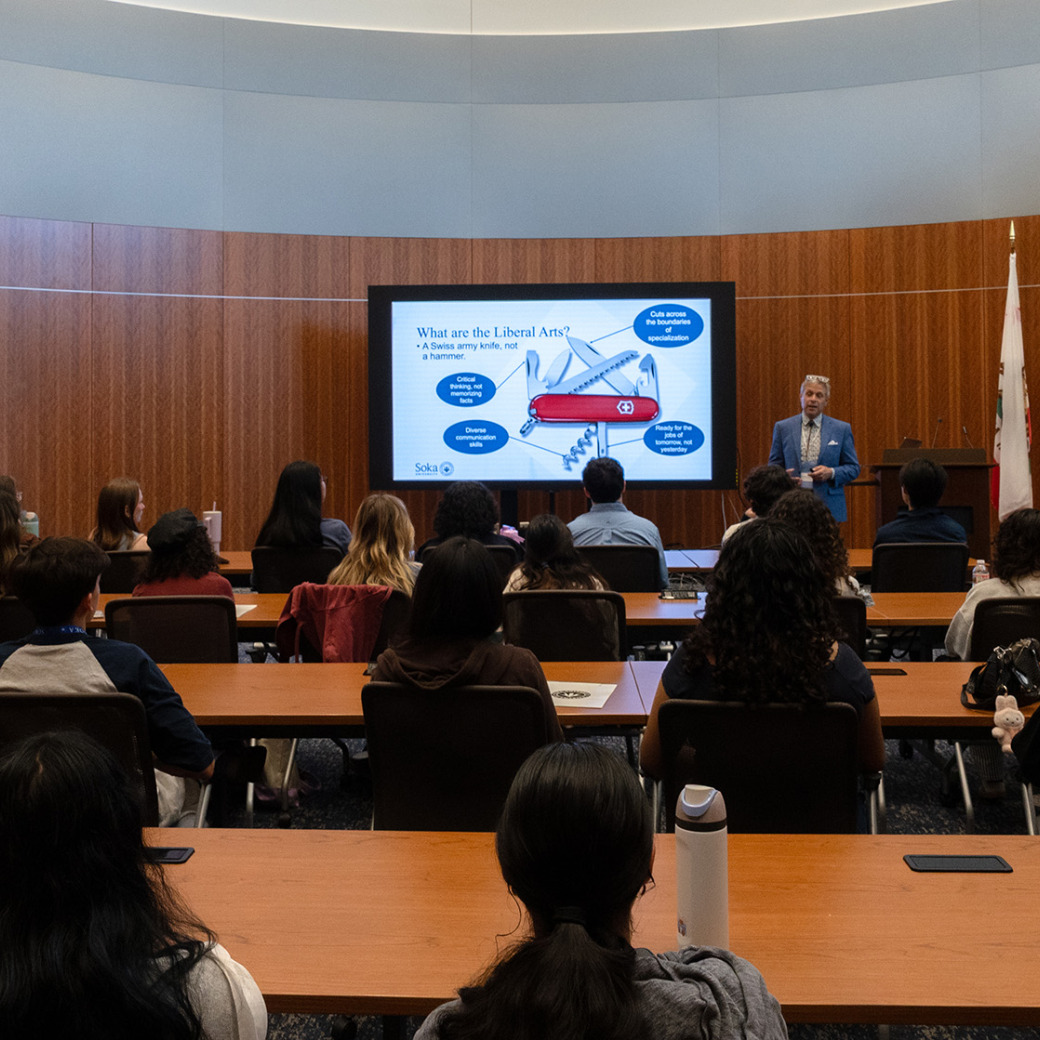 A presenter in a blue blazer stands before a lecture hall audience, speaking next to a large digital screen displaying a slide titled "What are the Liberal Arts?" featuring an illustration of a Swiss Army knife.