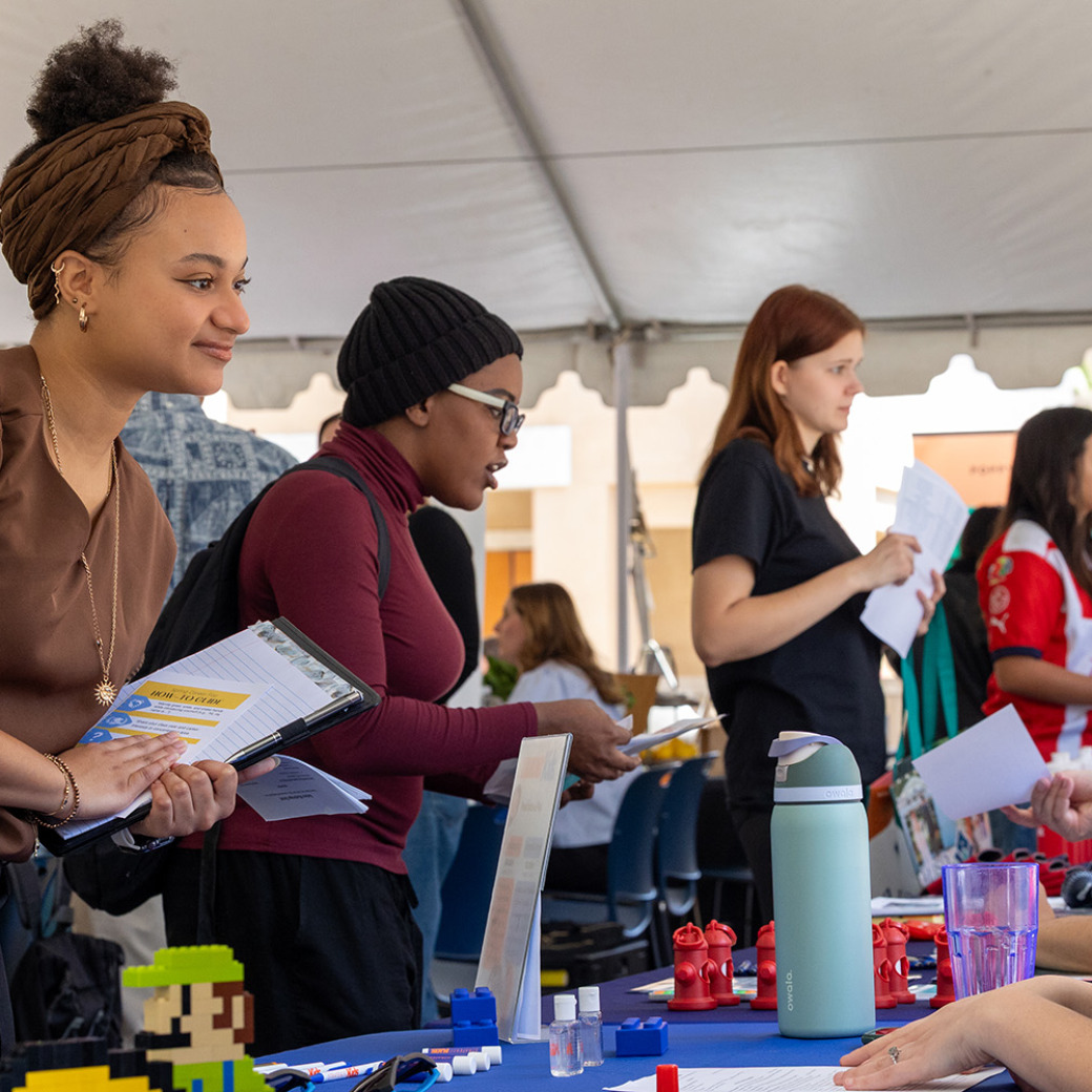 A young woman with a brown headwrap and gold jewelry smiles while leaning over a table at an outdoor career fair, holding a clipboard and documents. Other students and recruiters are visible under a large white tent in the background.