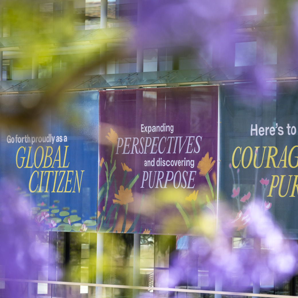 Marketing signs with mission oriented verbiage are posted on outside the PAC and photographed through purple jacaranda flowers.