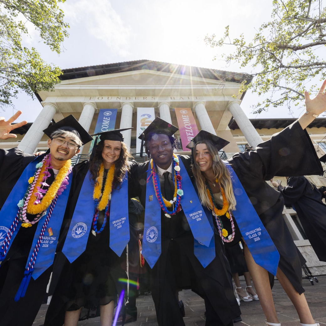 Four graduates wearing black gowns, royal blue sashes, and yellow cords, stand in front of Founders Hall pillars and look down at the camera.