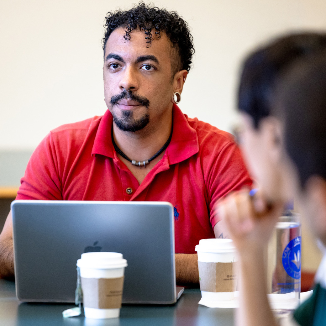 A man with curly hair and a red polo shirt listening attentively behind a laptop during a discussion.