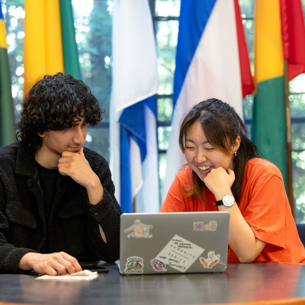A man with dark curly hair wearing a black shirt looks at a laptop with a dark-haired woman wearing an orange shirt. 