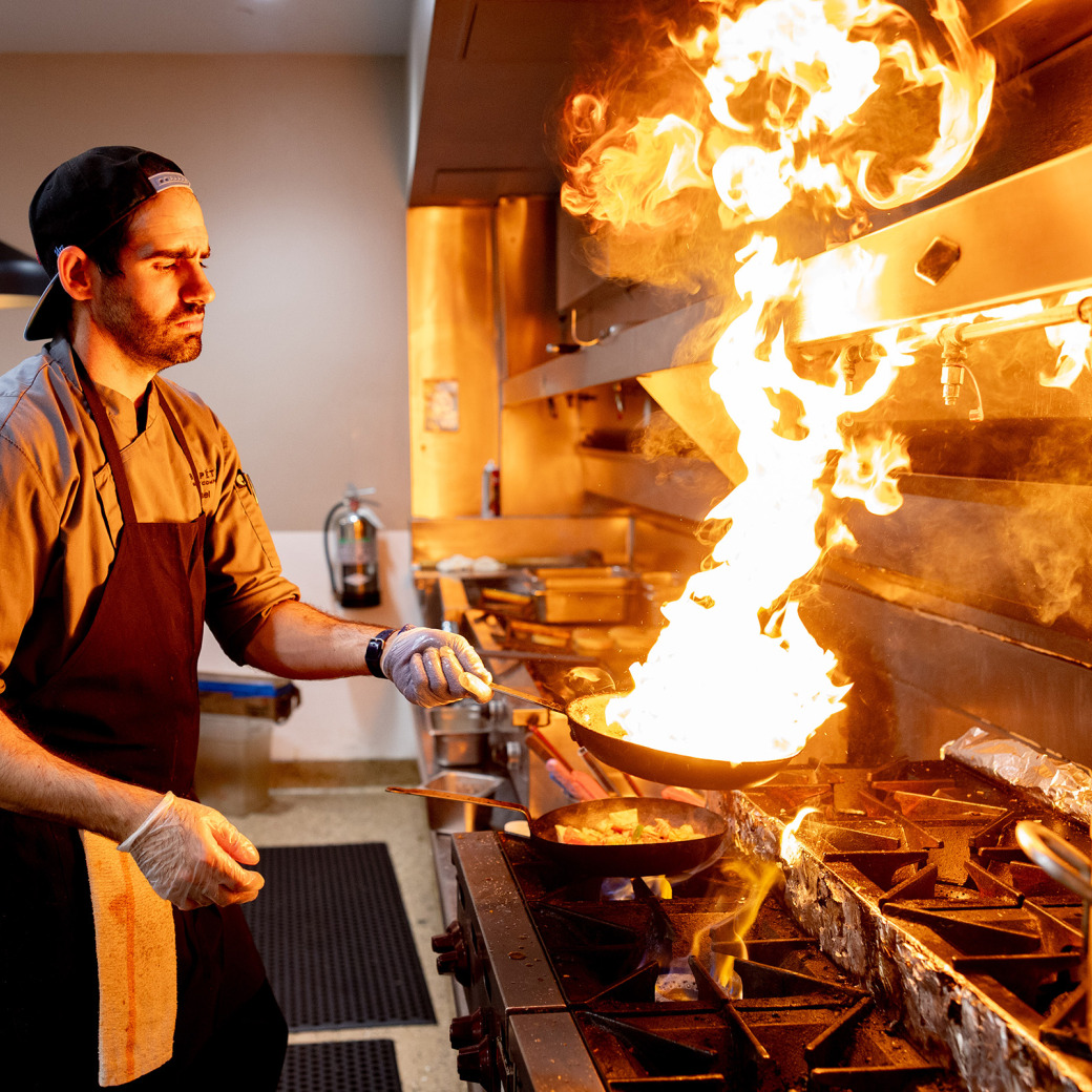 A male chef in a grey uniform and black apron intensely focuses on a large burst of fire erupting from a frying pan on a commercial stove. He wears a backwards baseball cap and gloves while tossing food in a professional kitchen setting.