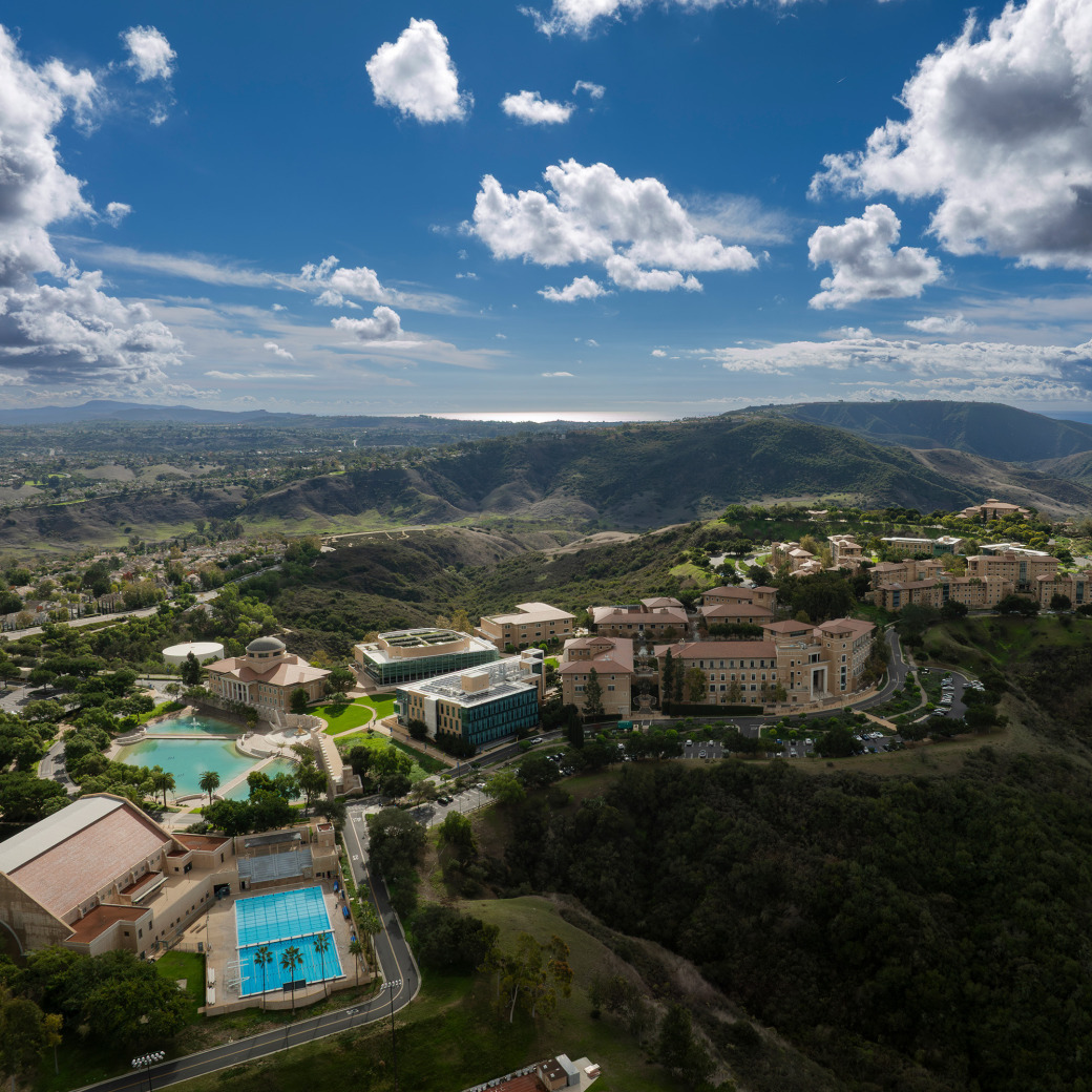 An aerial drone photograph of Soka University of America featuring a bright blue swimming pool, Peace Lake, and academic buildings nestled in rolling hills under a cloudy blue sky.