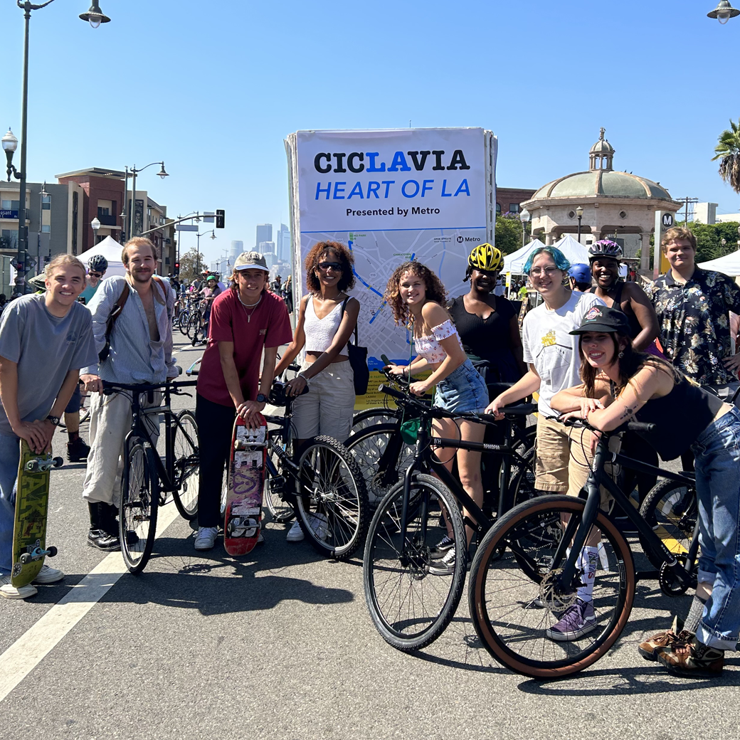 A large group of students holding skateboards and bicycles pose for a photo in downtown LA.