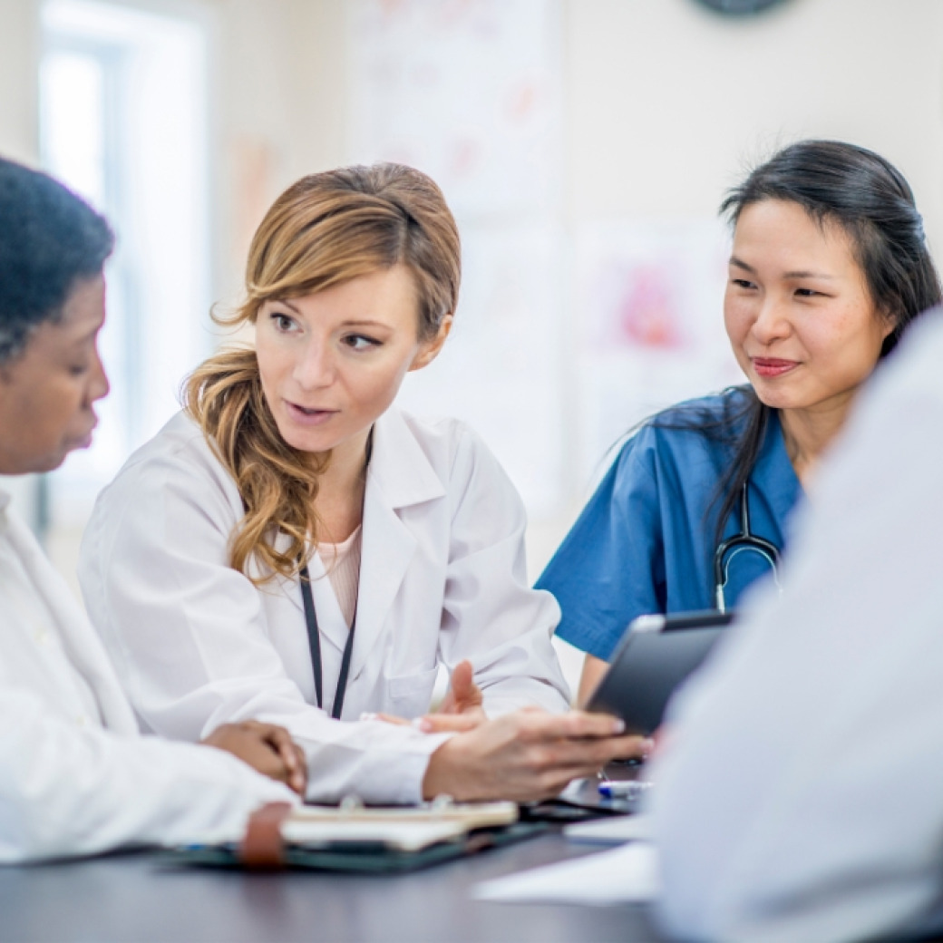 Four medical professionals wearing scrubs, white coats, and stethoscopes have a conversation.
