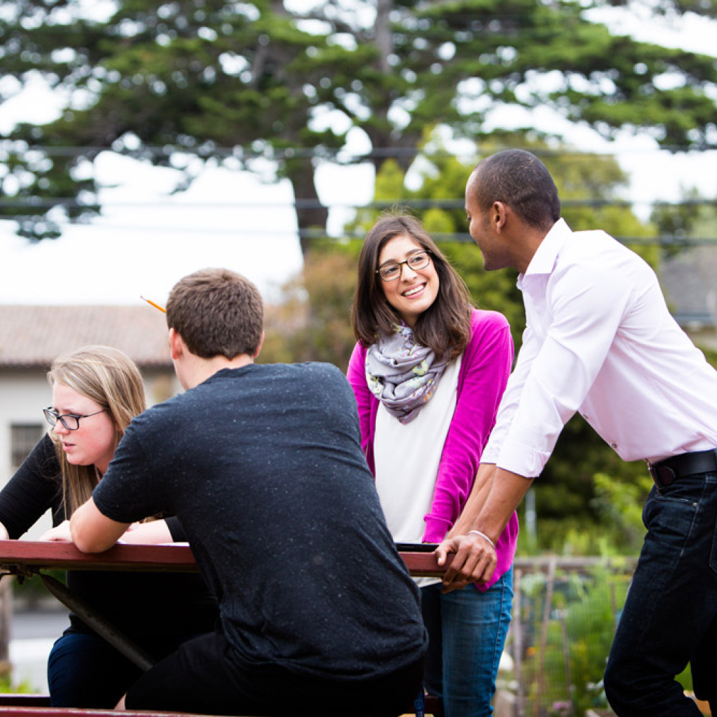 A woman with blonde hair and glasses is looking at a laptop with a man wearing a dark tshirt as two people stand at the end of a picnic table and chat.