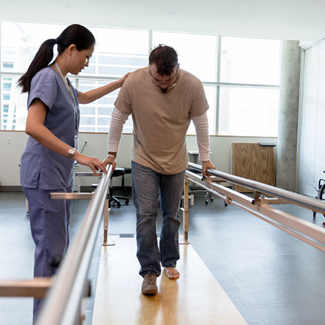 PT assisting a patient walking between parallel bars 