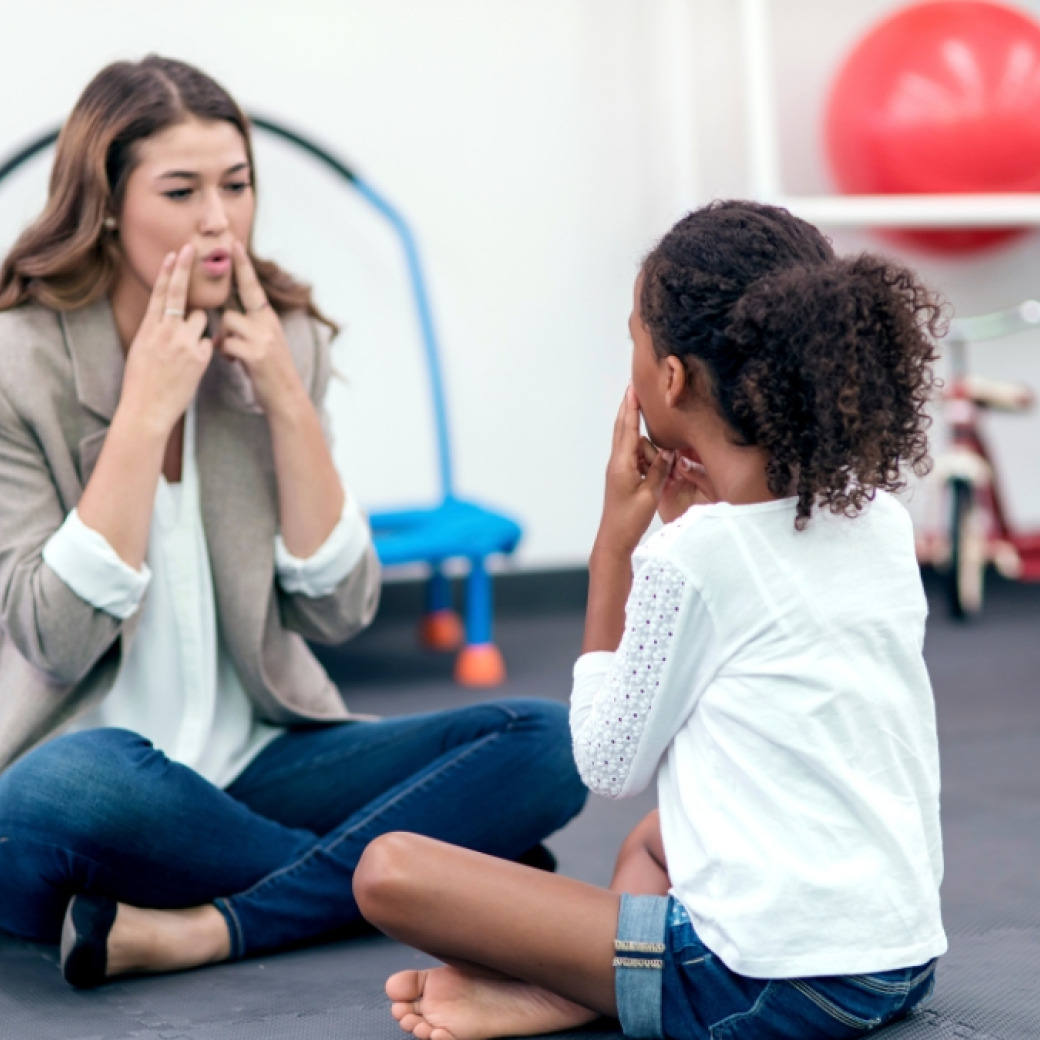 A speech-language pathologist demonstrating mouth and tongue positioning to a young child 