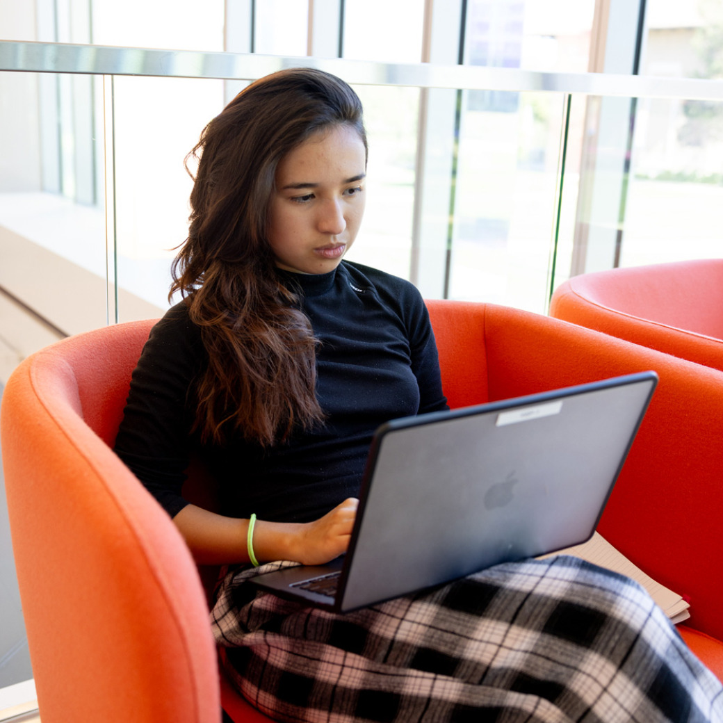 A student with brown hair wearing a black longsleeve and black and white checkered pants works on her laptop while sitting in a bright orange cushioned chair.