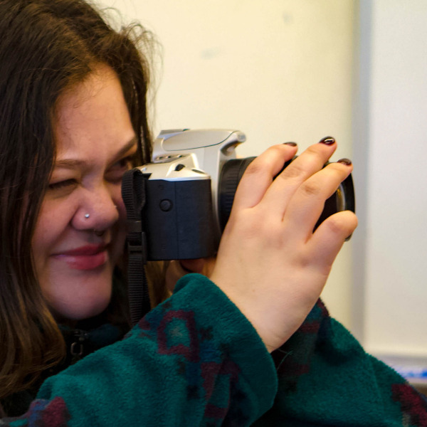 A smiling woman with dark hair and a nose piercing holds a silver and black DSLR camera up to her eye, preparing to take a photo. She is wearing a patterned teal fleece jacket.