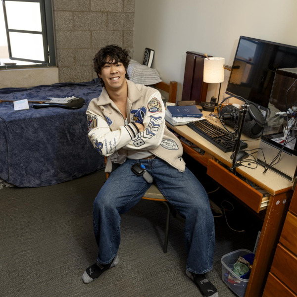 A male student with wavy brown hair wears a white zipup and blue jeans as he sits on a chair at his desk in the dorm room. His twin bed behind him has a blue blanket with an electric guitar laying on top of it. 