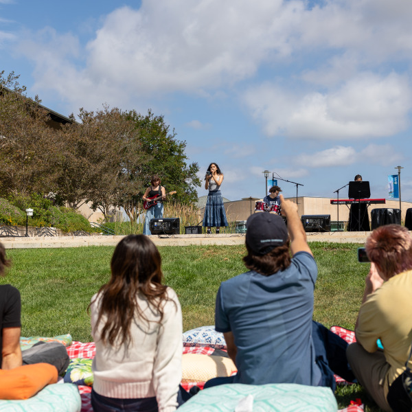 Four students cheer sit with their backs to the camera cheering on student performers on an outdoor stage.