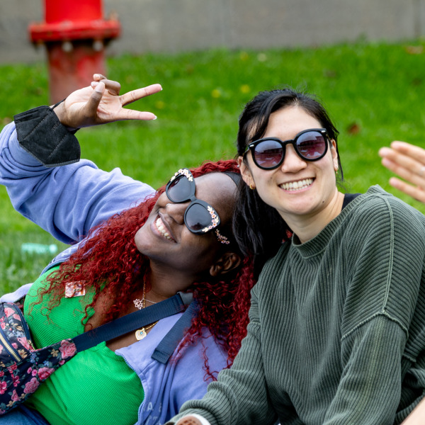 Two women wearing sunglasses pose for a photo. One is giving a peace sign.