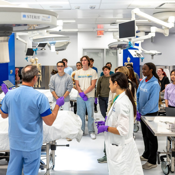 A group of students stands in a modern anatomy lab or surgical training facility, listening to an instructor in blue scrubs. Several lab stations with covered tables, medical lighting, and surgical tools are visible around the bright, sterile room.