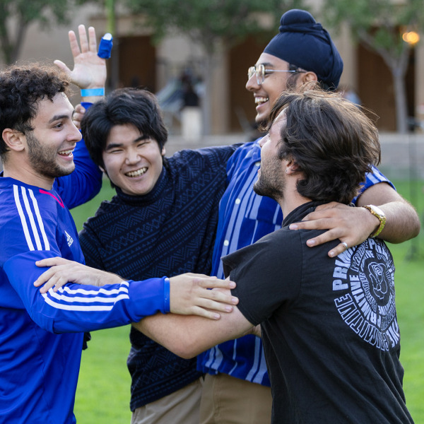  Four young men laughing and hugging on a grassy field.
