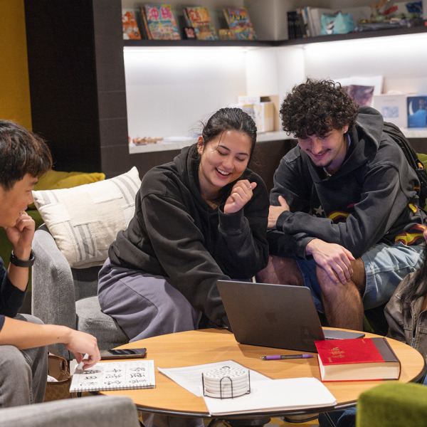 Four students smiling and collaborating around a laptop on a coffee table in a comfortable campus lounge setting.