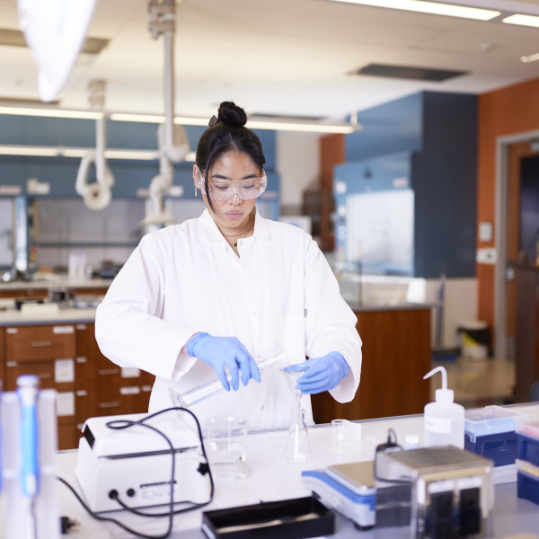 A female student with black tied-back hair wears a white lab coat, safety glasses, and blue gloves as she performs science experiments in a lab.