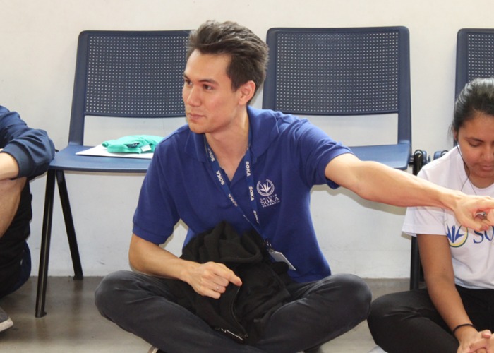 Scott Bower sits cross-legged on the floor in between two students and is pointing to his left