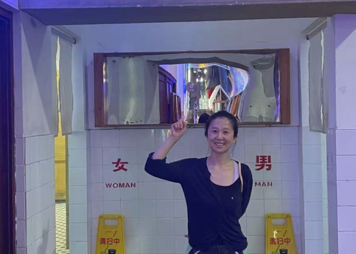 Dr. Yang Allen smiling and pointing upwards, standing in front of public restroom entrances with "WOMAN" and "MAN" signs.