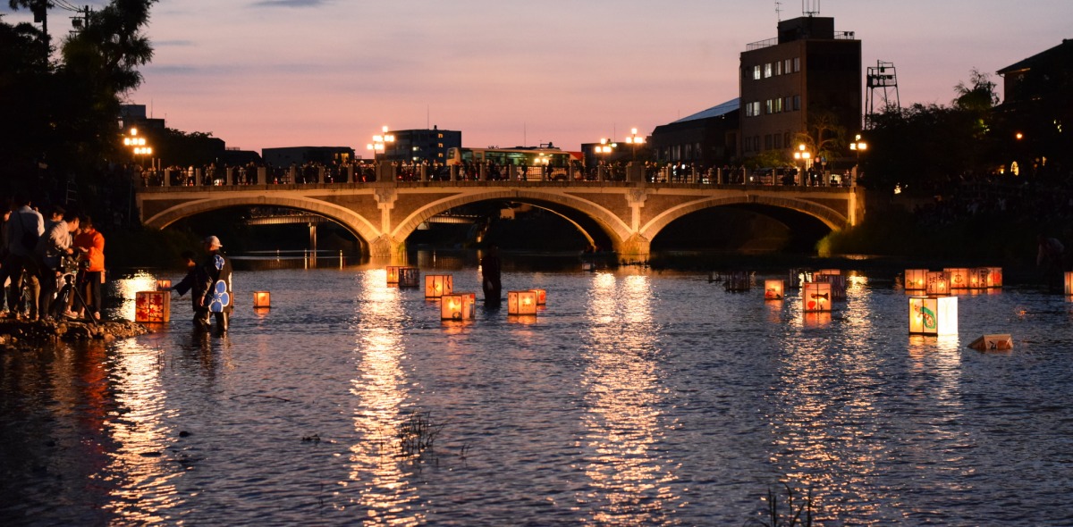 Picture of a bridge illuminated from behind by a sunset and underneath by lanterns floating on the water.