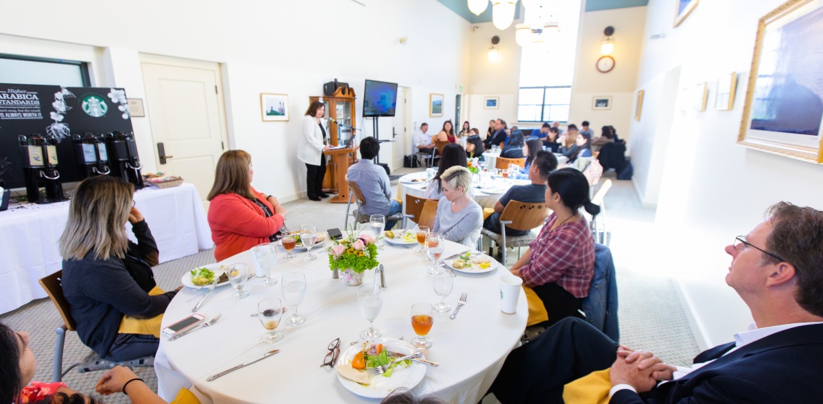 Image of a speaker at a podium addressing a group of people seated at tables.