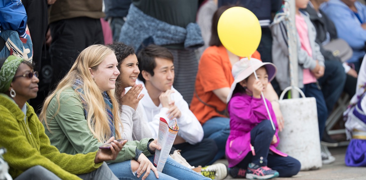 People at a festival sit on the ground while watching entertainment. One girl is holding a yellow balloon.